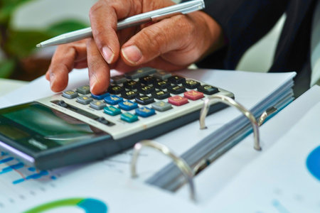 Close-up of hands calculating on a calculator and holding a pen over financial papers and a binder.の写真素材