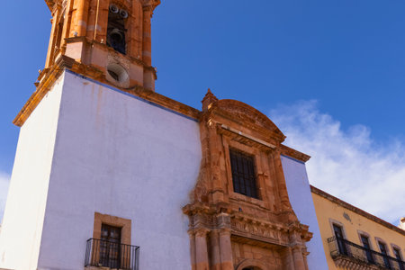 A white church building with a richly decorated orange facade and a tall bell tower stands under a vibrant blue sky with scattered clouds.の写真素材