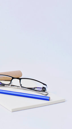 A pair of reading glasses rests on a blue folder with a pen lying on top. Minimalist composition with a light background.の写真素材