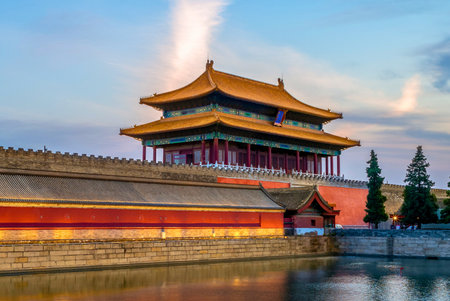 A majestic ancient Chinese imperial palace structure with ornate rooftops stands behind a red wall, its reflection shimmering in the calm water below.の写真素材