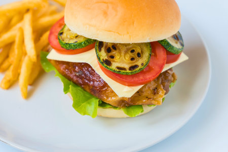A gourmet chicken burger stacked high with grilled zucchini, cheese, tomato, and lettuce, served with crispy french fries.の写真素材
