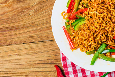 A close-up shot of a plate filled with savory noodles and colorful vegetables, presented on a wooden surface with a checkered cloth.の写真素材