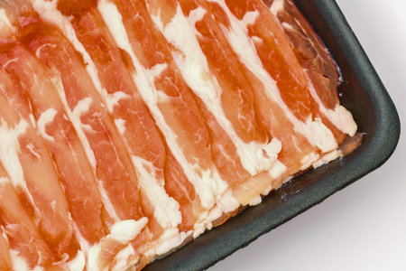 Close-up overhead view of uncooked bacon strips neatly arranged in a black tray, showcasing the red meat and white fat marbling.の写真素材