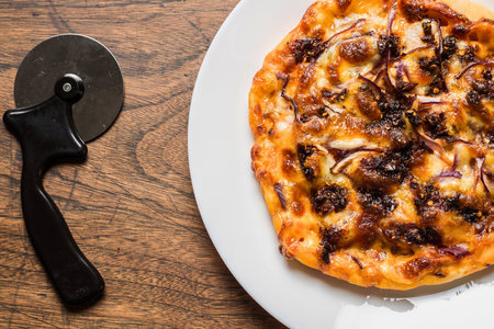 A close-up overhead shot of a freshly baked pizza on a white plate next to a pizza cutter on a wooden table.の写真素材