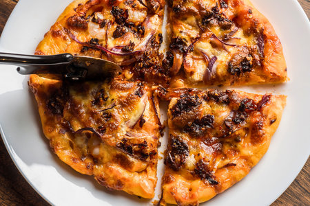 A close-up overhead view of a golden-brown flatbread pizza topped with melted cheese and sweet caramelized onions, being served with tongs.の写真素材