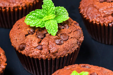 Close-up view of rich chocolate chip muffins, one prominently garnished with vibrant green mint leaves, against a dark background.の写真素材