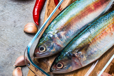 Close-up overhead view of two vibrant, iridescent fish on a wooden cutting board with red chili peppers and garlic cloves.の写真素材
