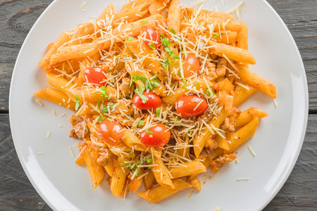 A close-up overhead view of a white oval plate filled with penne pasta, topped with a rich sauce, cherry tomatoes, and shredded cheese.の写真素材