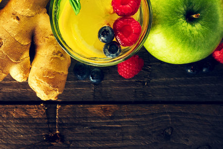 A vibrant yellow smoothie in a glass, garnished with berries and a green straw, sits beside fresh ginger and a green apple on a wooden surface.の写真素材