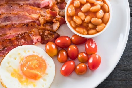 A close-up of a delicious breakfast featuring crispy bacon, a soft-boiled egg, baked beans, and fresh cherry tomatoes on a white plate.の写真素材