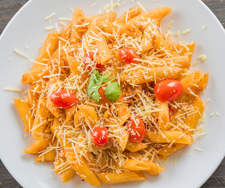 A close-up overhead view of a white plate filled with penne pasta, topped with a rich sauce, cherry tomatoes, and grated cheese.の写真素材