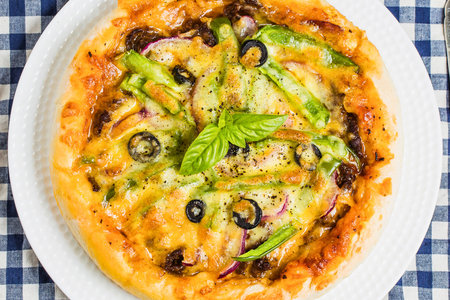 A close-up overhead view of a golden-brown vegetable pizza with melted cheese, olives, peppers, and fresh basil.の写真素材