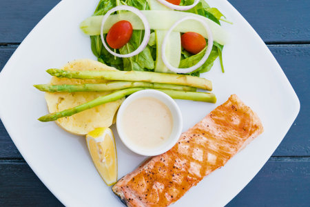A beautifully plated meal featuring grilled salmon, steamed asparagus, a fresh salad, and a creamy dipping sauce, presented on a white plate.の写真素材
