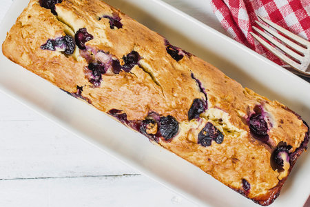 A close-up overhead shot of a freshly baked blueberry loaf cake on a white platter, with a fork and red checkered napkin in the background.の写真素材