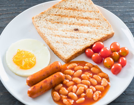 A hearty breakfast featuring grilled toast, sausages, baked beans, a fried egg, and cherry tomatoes on a white plate.の写真素材