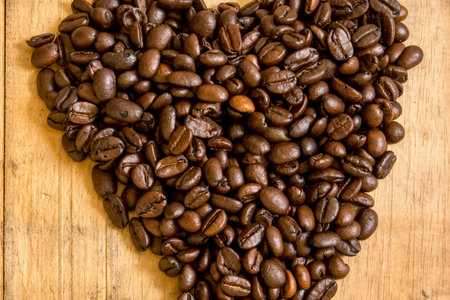 A close-up overhead view of dark roasted coffee beans artfully arranged into a heart shape on a light wooden background.の写真素材