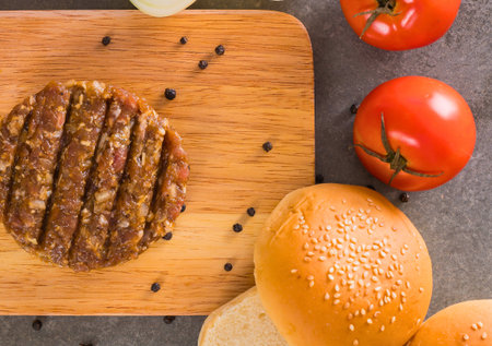 A juicy, grill-marked burger patty sits on a wooden board with tomatoes and burger buns, ready for assembly.の写真素材