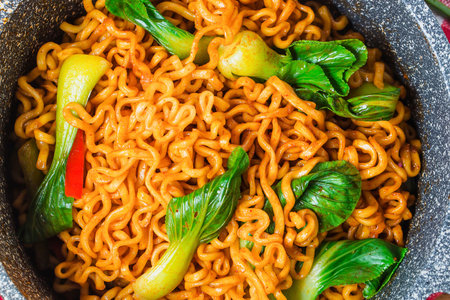 A close-up overhead view of a bowl filled with vibrant, spicy instant noodles, garnished with fresh green bok choy and red chili peppers.の写真素材