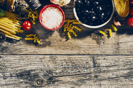 A top-down view of raw pasta, tomatoes, garlic, salt, and olives arranged on a weathered wooden surface, ready for cooking.の写真素材