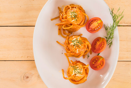 Three artfully arranged pasta nests coated in a vibrant orange sauce, garnished with fresh herbs and served with halved cherry tomatoes.の写真素材