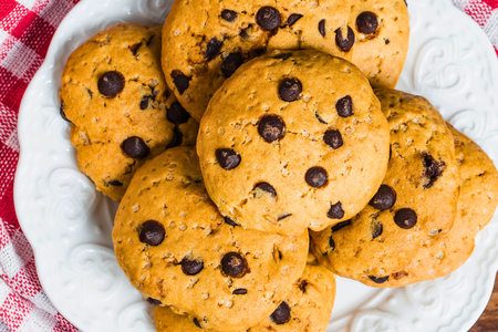 A close-up overhead view of a stack of golden brown chocolate chip cookies, generously studded with dark chocolate chips, resting on a white plate.の写真素材