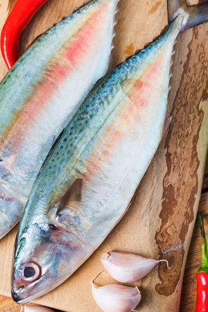 Two whole, uncooked fish with vibrant scales lie on a wooden board alongside chili peppers and garlic cloves.の写真素材