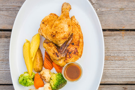 A perfectly roasted chicken sits on a white oval plate with a side of steamed vegetables and a small dish of sauce.の写真素材