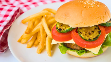 A close-up of a juicy cheeseburger with fresh toppings and a side of golden french fries, presented on a white plate with a red and white checkered napkin.の写真素材