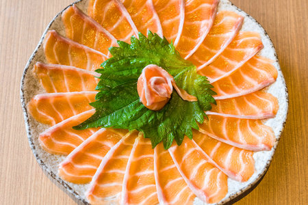A platter of vibrant salmon sashimi is artfully arranged in a circular pattern, garnished with fresh green leaves and a dollop of ginger.の写真素材