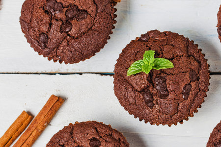 Close-up overhead view of rich chocolate muffins, one garnished with fresh mint, alongside cinnamon sticks on a white wooden surface.の写真素材