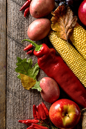 A vibrant arrangement of fresh fall produce including corn, peppers, apples, potatoes, and chili peppers rests on a weathered wooden surface.の写真素材