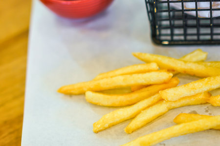 Close-up of crispy golden french fries scattered on a white surface next to a red dipping sauce and a black wire basket.の写真素材