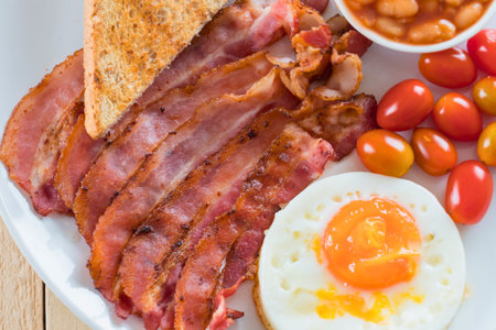 A hearty and delicious full English breakfast featuring crispy bacon, a fried egg, toast, cherry tomatoes, and baked beans.の写真素材