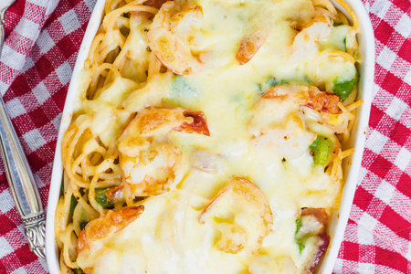 A close-up overhead view of a white oval baking dish filled with spaghetti, shrimp, and melted cheese, set on a red and white checkered cloth.の写真素材