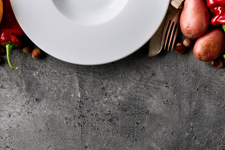 A white plate sits on a textured gray surface, surrounded by a vibrant assortment of fresh vegetables and potatoes.の写真素材