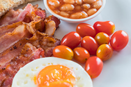 A close-up shot of a hearty full English breakfast featuring crispy bacon, baked beans, a fried egg, and fresh cherry tomatoes.の写真素材