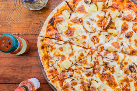 A close-up aerial view of a freshly baked Hawaiian pizza, featuring pineapple and ham, on a rustic wooden surface.の写真素材