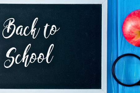 A chalkboard with 'Back to School' written in cursive, next to an apple and a pair of glasses on a blue wooden surface.の写真素材