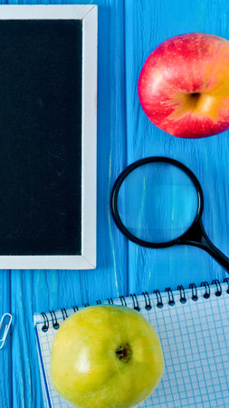 A tablet, magnifying glass, notebook, and two fruits are arranged on a vibrant blue wooden surface,...
