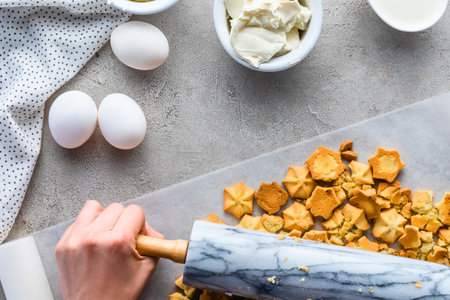 Overhead view of baking ingredients including eggs and a bowl of something creamy, with a hand using a rolling pin on parchment paper.の写真素材