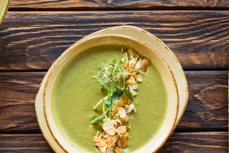 A creamy green soup is presented in an artisanal bowl, garnished with delicate herbs and crispy toppings, set against a textured wooden background.の写真素材