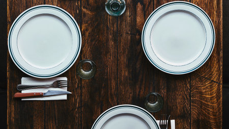A top-down view of a rustic wooden table set with three white oval plates, silverware, and napkins, ready for a meal.の写真素材