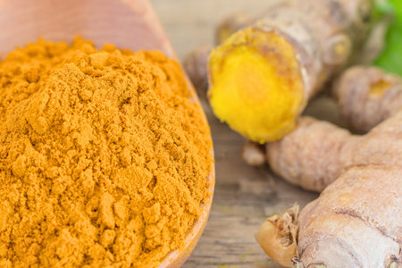 Close-up of bright yellow turmeric powder in a wooden scoop next to fresh turmeric rhizomes on a rustic wooden background.の写真素材