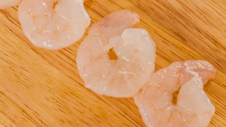 Three raw shrimp tails are arranged diagonally on a warm-toned wooden surface, highlighting their translucent pinkish-white color and delicate texture.の写真素材