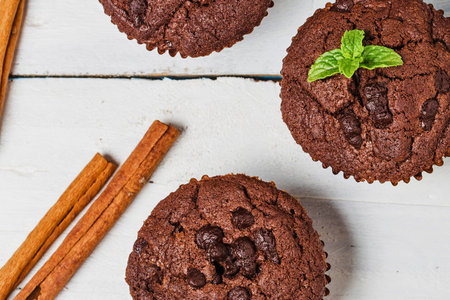 Rich chocolate muffins studded with chocolate chips, garnished with mint, and served with cinnamon sticks on a white wooden background.の写真素材