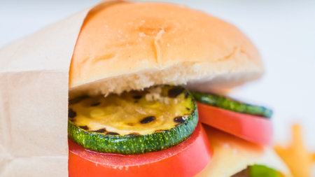 A close-up, slightly blurred shot of a vegetarian burger featuring grilled zucchini, tomato slices, and a soft bun, wrapped in paper.の写真素材