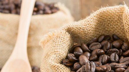Close-up of burlap sacks filled with dark roasted coffee beans, with a wooden spoon in the foreground, suggesting freshness and preparation.の写真素材