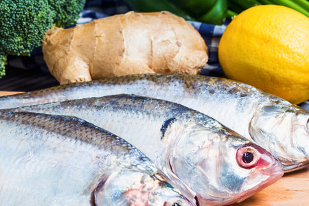 Three fresh whole fish are arranged with broccoli, ginger, and a lemon, suggesting a healthy and flavorful meal preparation.の写真素材