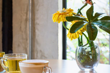 A close-up of a table with a latte and a vase of yellow flowers by a window.の写真素材