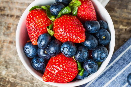 A close-up overhead view of a white bowl filled with vibrant red strawberries and plump blue blueberries, presented on a rustic wooden surface.の写真素材